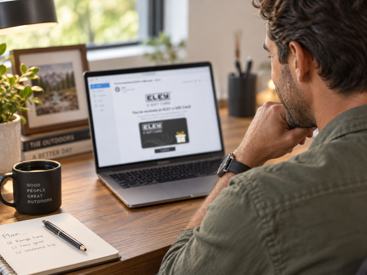 A man sits at a wooden desk looking at a laptop screen displaying an ELEY e-Gift Card he received, with a coffee mug, notebook, and framed picture on the desk.